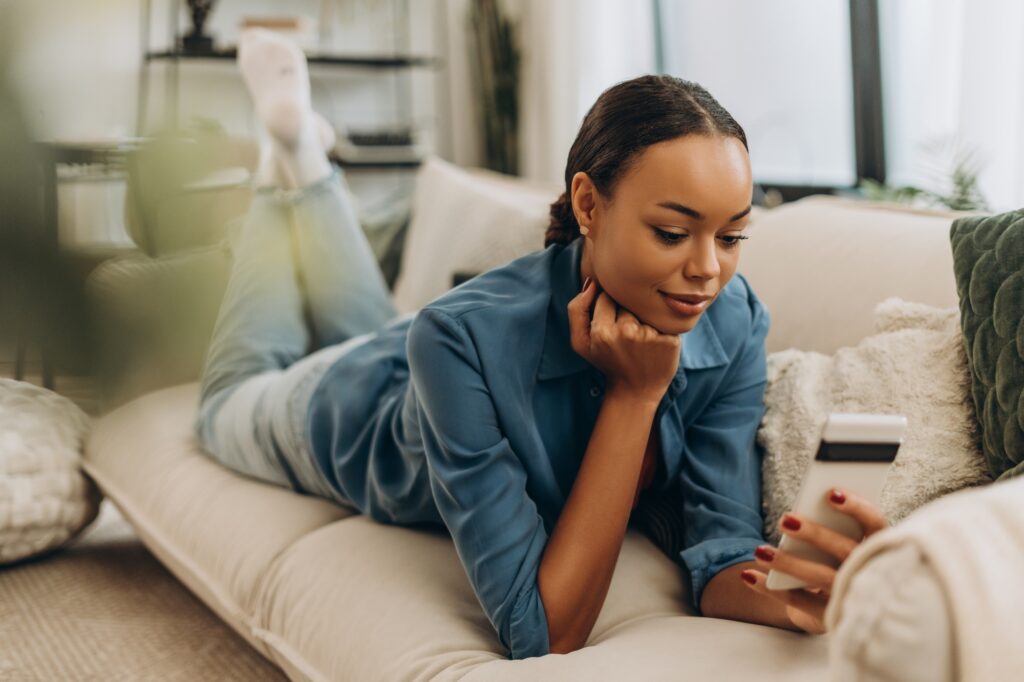 Smiling, African American woman holding mobile phone, social media lying on comfortable sofa