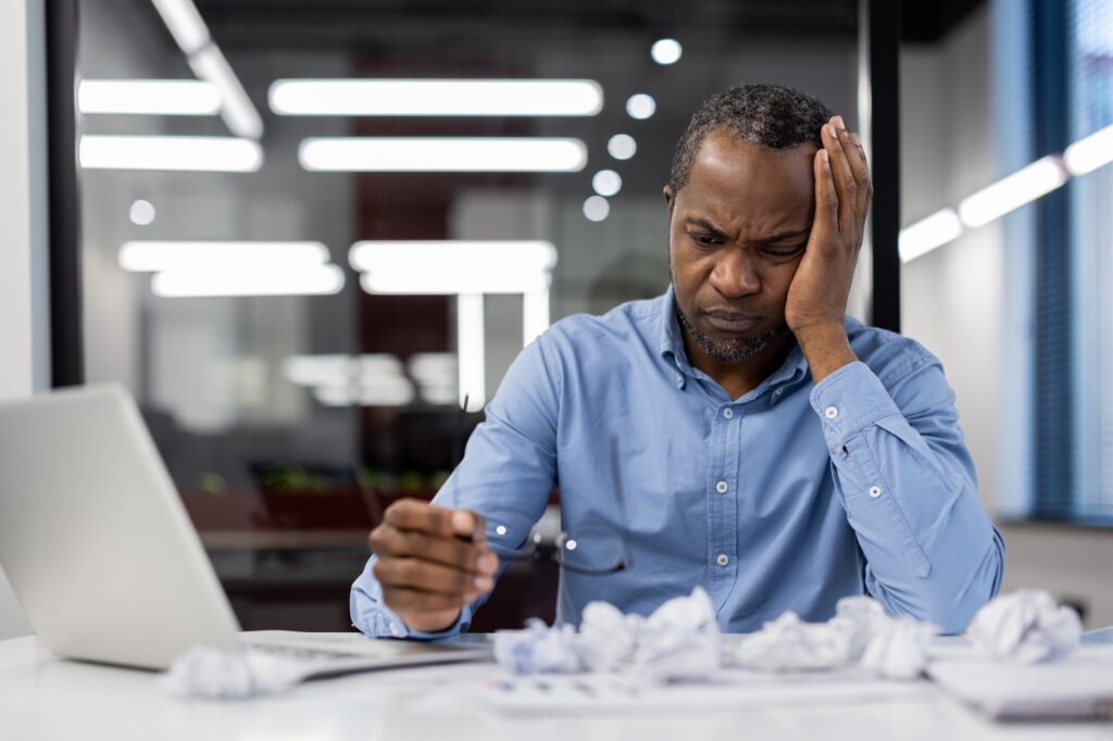 Stressed office worker dealing with frustration at desk surrounded by crumpled papers. Overwhelmed