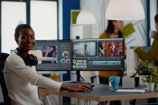 African video editor looking at camera smiling editing video project in post production software working in creative studio office. Videographer editing audio film montage on professional computer